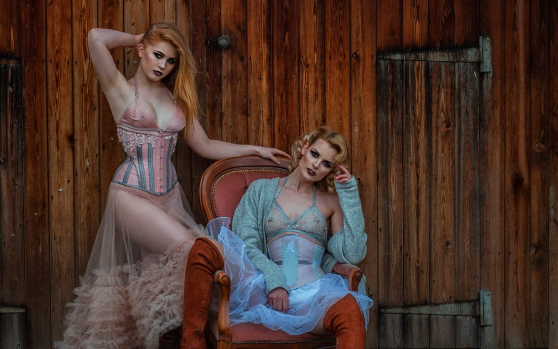 Two models wear pink and grey corsets in front of a barn door