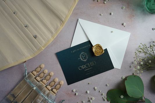 A teal and gold gift card with wax sealed envelope alongside an corset and suspenders.